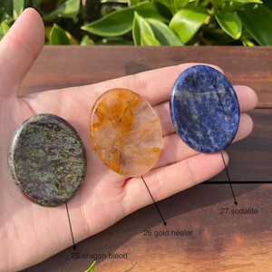 May include: Three smooth, polished oval-shaped stones. The stone on the left is green with red and black swirls. The middle stone is clear with gold flecks. The stone on the right is blue with white streaks. The stones are labeled "25. dragon blood", "26. gold healer", and "27. sodalite".