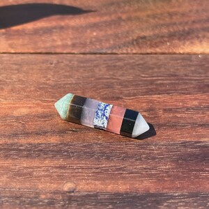 May include: A multi-colored crystal wand with seven different colored stones. The wand is pointed at one end and has a smooth, polished surface.
