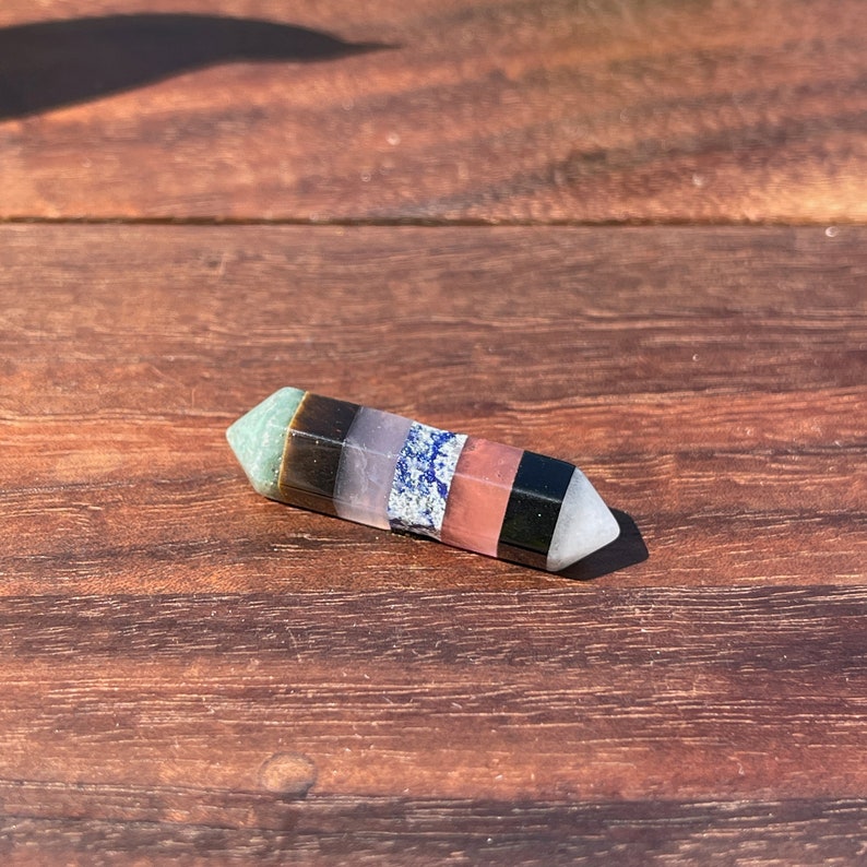 May include: A multi-colored crystal wand with seven different colored stones. The wand is pointed at one end and has a smooth, polished surface.