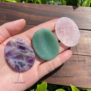 May include: Three oval-shaped polished stones, amethyst, green aventurine, and rose quartz, are displayed on a hand. The stones are labeled with their names.