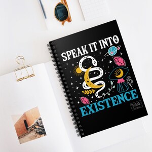Puede incluir: Cuaderno espiral negro con el texto "Speak it into existence" en letras blancas. El cuaderno tiene un diseño colorido con una serpiente, una luna, estrellas y cristales.