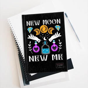 May include: Black journal with the text "New Moon New Me" in white lettering. The cover features a crescent moon, three moons in a circle, stars, and two hands holding a bottle of blue liquid. There are also two purple bottles with hearts on them.