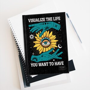 Puede incluir: Diario negro con un girasol y dos manos formando un círculo a su alrededor. El texto "Visualize the life you want to have" está impreso en la portada. El diario tiene un fondo negro con un diseño azul y amarillo.