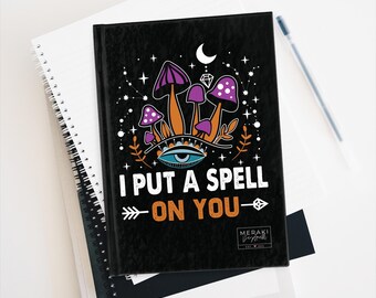 May include: Black journal with a colorful illustration of mushrooms, a crescent moon, stars, and an eye. The text "I put a spell on you" is written in orange and white. The journal has a black spine and a white logo that says "MERAKI" with a small design.