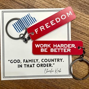 May include: Two red keychains with white text reading "FREEDOM" and "WORK HARDER, BE BETTER." A card with the quote "GOD, FAMILY, COUNTRY. IN THAT ORDER." by Charlie Kirk is also present. The keychains are attached to metal rings.