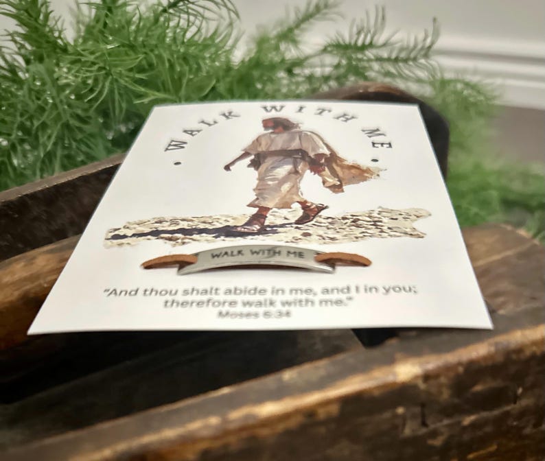 May include: A white card with the words "WALK WITH ME" and an illustration of a person walking. The card also features a quote from Moses 6:34. A small metal accent with the words "WALK WITH ME" is below the image.