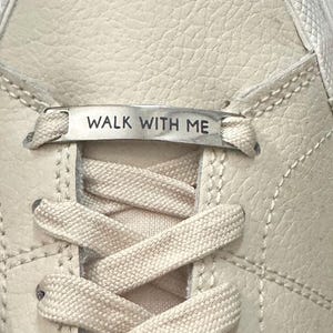 May include: Close-up of a cream-colored sneaker with a textured leather upper. The laces are white, and a silver tag across the laces reads "WALK WITH ME". The shoe has visible stitching details.