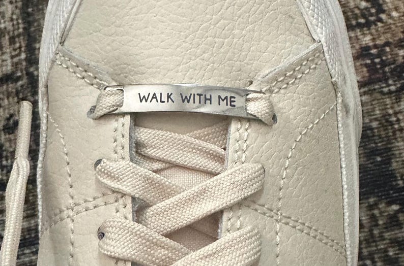 May include: Close-up of a cream-colored sneaker with a textured leather upper. The laces are white, and a silver tag across the laces reads "WALK WITH ME". The shoe has visible stitching details.