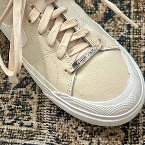 May include: Cream-colored sneakers with white soles and matching laces. The shoe features a textured leather upper and a metal tag that reads "WALK WITH ME". The design is minimalist and modern, suitable for casual wear.