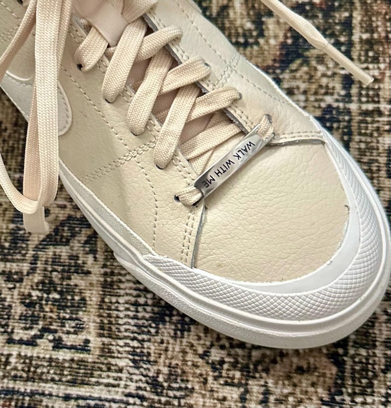 May include: Cream-colored sneakers with white soles and matching laces. The shoe features a textured leather upper and a metal tag that reads "WALK WITH ME". The design is minimalist and modern, suitable for casual wear.