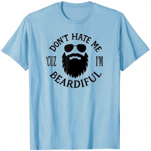 Puede incluir: Camiseta azul claro con un gráfico negro de una barba con gafas de sol y el texto "DON'T HATE ME 'CUZ I'M BEARDIFUL" en un diseño circular. La camiseta está hecha de un material suave.