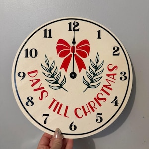 May include: A round wooden Christmas countdown clock with a cream-coloured face. The clock features black numerals, a red bow, green sprigs, and the words "DAYS TILL CHRISTMAS" in red. The clock is held by a person.