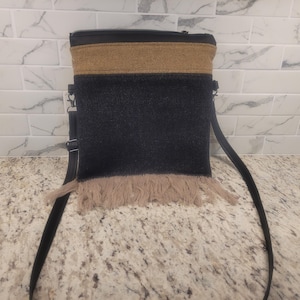 May include: A black crossbody bag with a tan fringe and a tan band across the top. The bag has a black zipper and a black adjustable strap. The bag is made of a soft material.