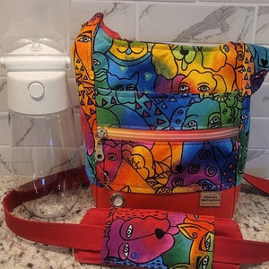 May include: A red crossbody bag with a colorful, abstract print of faces and swirls. The bag has a zippered pocket and a detachable strap. The bag is made by "Whimsical Making New".