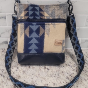May include: A blue and beige patterned crossbody bag with a black leather bottom and a blue, black, and gray patterned strap. The bag has a zipper closure and a brown leather tag that says "Whatcha Making Now".