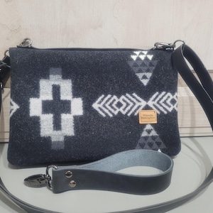 May include: A dark gray felt crossbody bag featuring a white and light gray geometric pattern. The bag includes a black leather shoulder strap and a detachable black leather wrist strap. A small rectangular tan label reads "Whertha Makington".