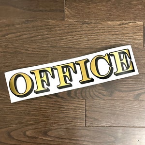 May include: Gold "OFFICE" sign with black outline on a white background. The sign is on a dark wood floor.
