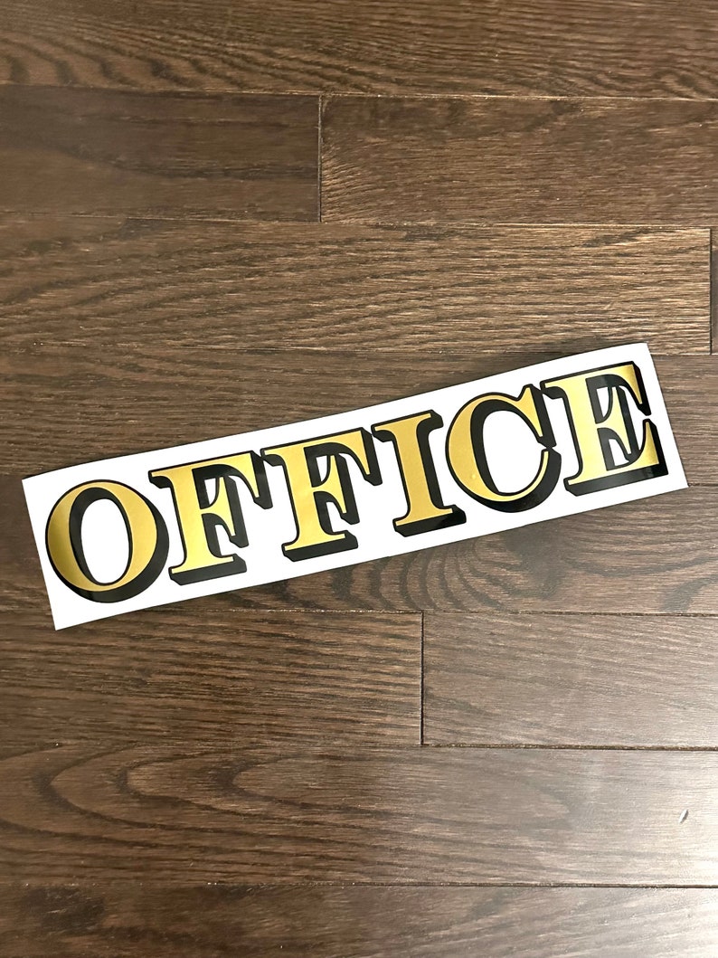 May include: Gold "OFFICE" sign with black outline on a white background. The sign is on a dark wood floor.