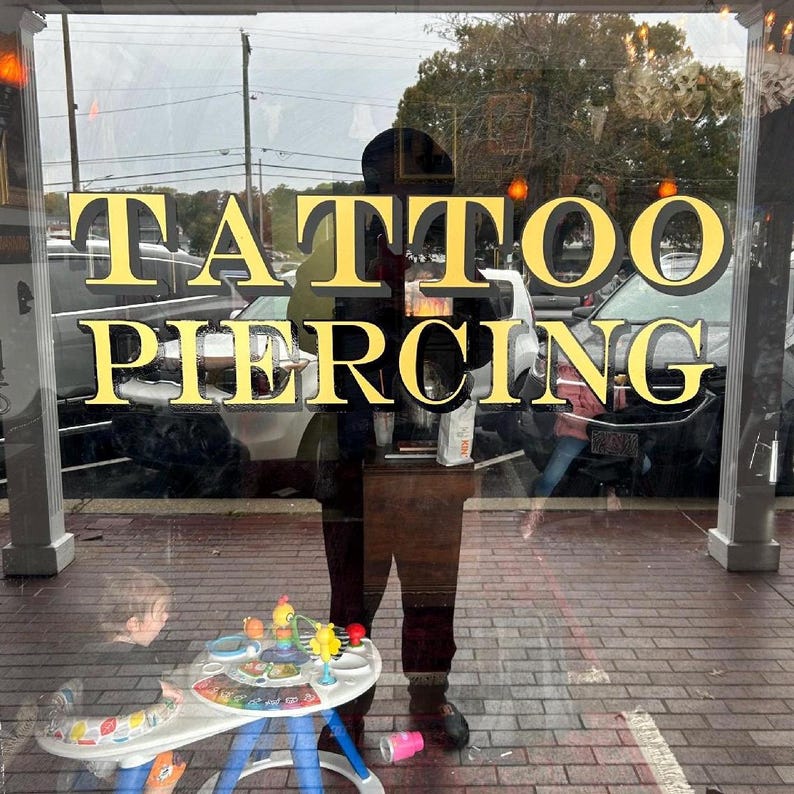 May include: A storefront window with the words "TATTOO PIERCING" in gold lettering. A person is reflected in the window, and a child is playing with a toy in the foreground.