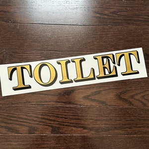 May include: A white sign with the word "TOILET" printed in gold letters with black outlines.