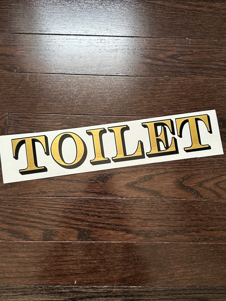 May include: A white sign with the word "TOILET" printed in gold letters with black outlines.