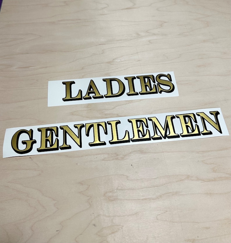 May include: Two white signs with black borders and gold lettering. The top sign reads "LADIES" and the bottom sign reads "GENTLEMEN".