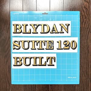 May include: Three white rectangular stickers with black outlines and gold lettering. The stickers read "BLYDAN", "SUITE 120", and "BUILT". The stickers are on a blue grid background.