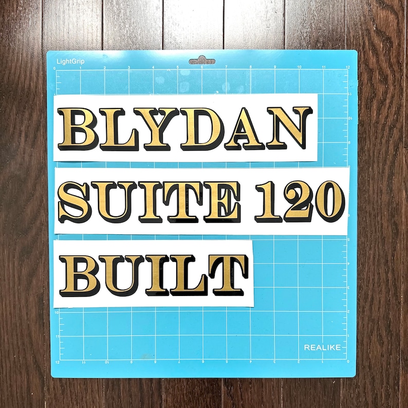 May include: Three white rectangular stickers with black outlines and gold lettering. The stickers read "BLYDAN", "SUITE 120", and "BUILT". The stickers are on a blue grid background.