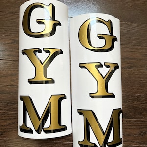 May include: Two white vinyl decals with gold lettering spelling out the word "GYM".