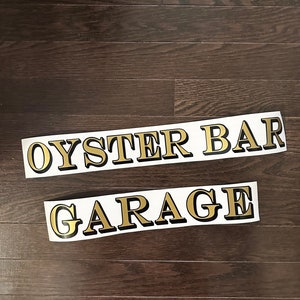 May include: Two gold vinyl stickers with black outlines. The top sticker says "OYSTER BAR" and the bottom sticker says "GARAGE".