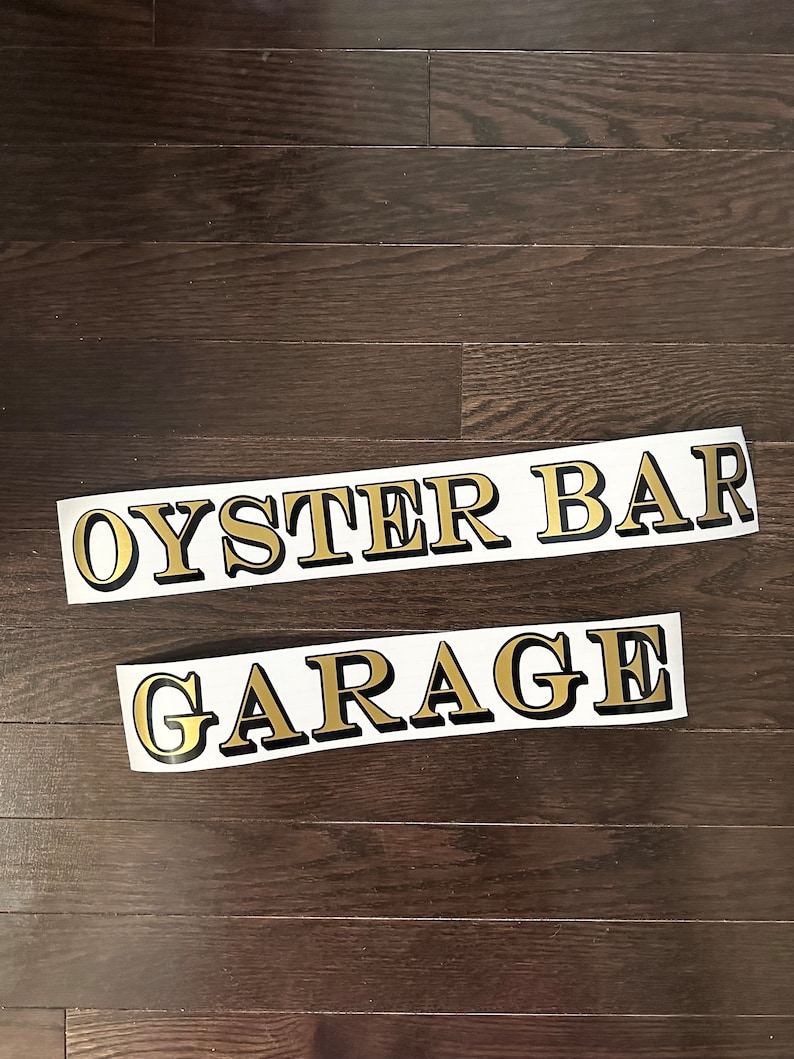 May include: Two gold vinyl stickers with black outlines. The top sticker says "OYSTER BAR" and the bottom sticker says "GARAGE".