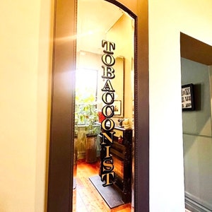 May include: A mirror reflection of a shop sign that reads "TOBACCONIST" in black letters with gold outlines. The sign is hanging on a wall inside a shop.