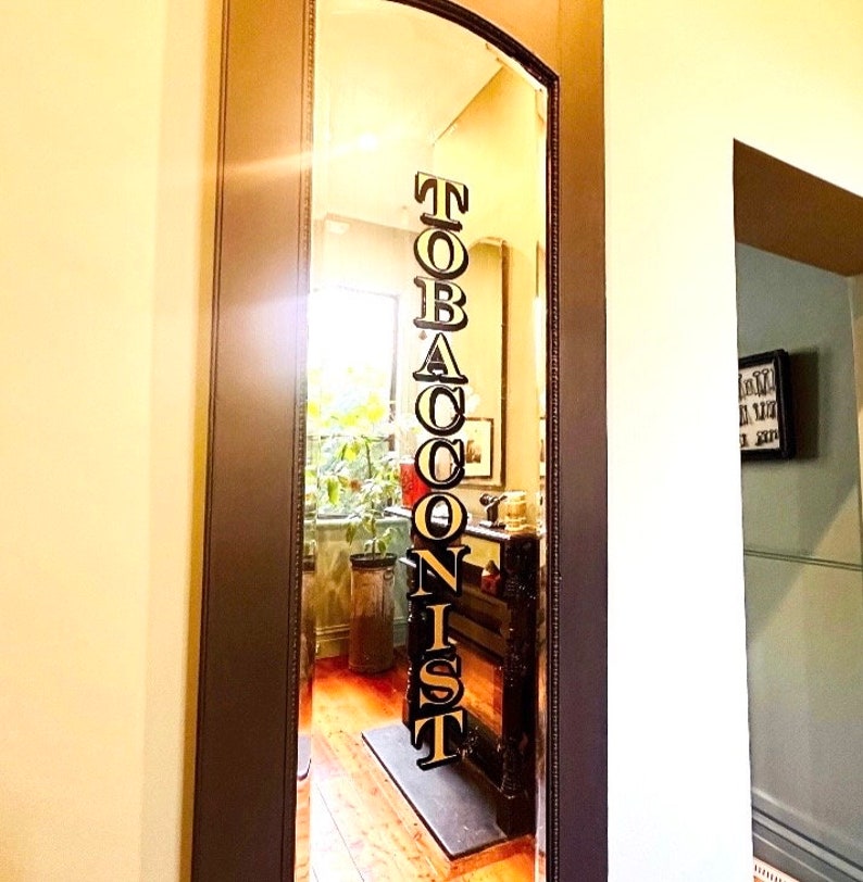 May include: A mirror reflection of a shop sign that reads "TOBACCONIST" in black letters with gold outlines. The sign is hanging on a wall inside a shop.