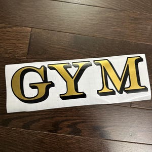 May include: A white sticker with the word "GYM" in gold letters with a black outline. The sticker is on a dark brown wooden surface.