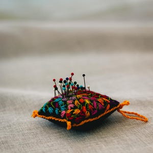 Könnte beinhalten: Ein quadratisches, schwarzes Nadelkissen mit bunten, gestickten Blumenmustern und einer leuchtend orangefarbenen Bordüre. Das Kissen ist mit verschiedenen Nadeln mit bunten Köpfen gefüllt. An der Seite ist eine orangefarbene Schlaufe angebracht.