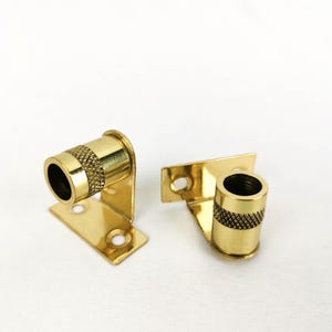 May include: Two gold metal curtain rod brackets with a decorative knurled design. The brackets have a flat base with two holes for mounting.