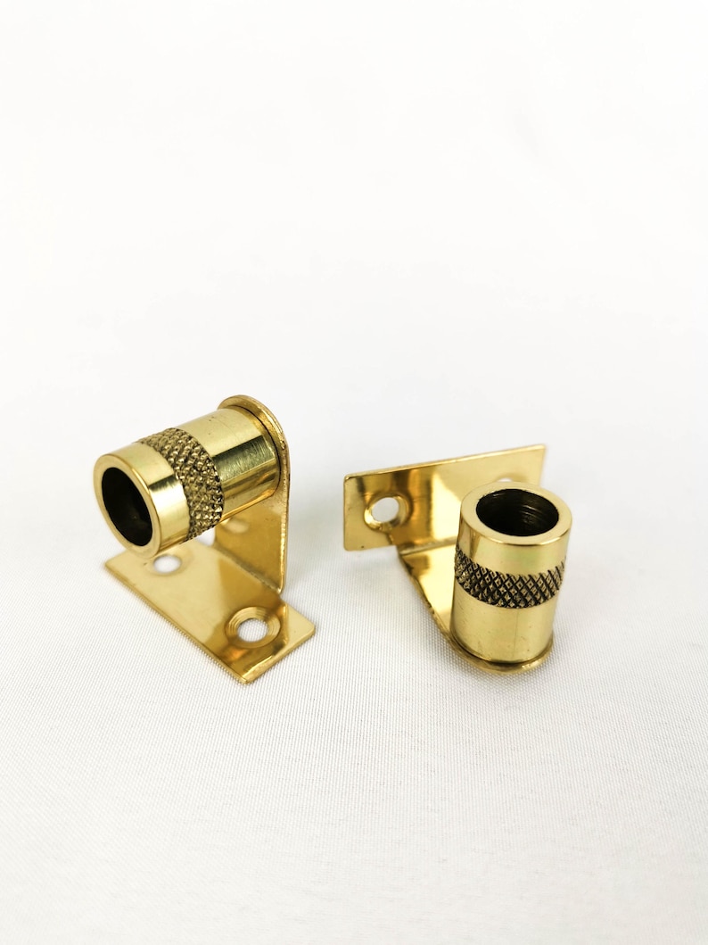 May include: Two gold metal curtain rod brackets with a decorative knurled design. The brackets have a flat base with two holes for mounting.