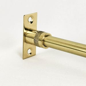 Op de afbeelding: Een gouden metalen gordijnroede met een vierkante wandbevestiging. De roede heeft een geribbelde knop om de spanning aan te passen.