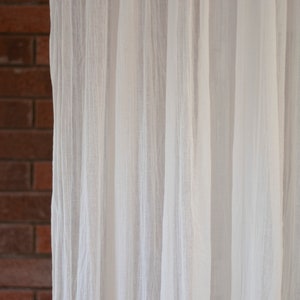 May include: Off-white, semi-sheer curtains hanging in vertical folds. The curtains are positioned in front of a red brick wall. The fabric appears to be lightweight and textured, allowing light to filter through.