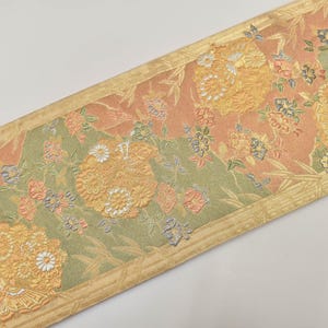 Può includere: Una striscia di tessuto decorativa con un motivo floreale. Il design presenta grandi fiori gialli, fiori più piccoli e fogliame in tonalità pesca, verde e oro. I bordi sono rifiniti con un bordo dorato.