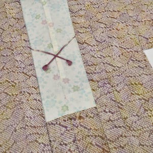 Puede incluir: Un kimono vintage con un diseño estampado en tonos morados, amarillos y marrones. El kimono presenta un panel blanco con motivos florales y una corbata decorativa. La tela tiene una apariencia texturizada.