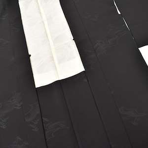 Könnte beinhalten: Ein schwarzer Kimono mit einem dezenten, tonalen Jacquardmuster aus stilisierten Wellen und Wolken auf dem gesamten Stoff. Das Kleidungsstück verfügt über ein kontrastierendes weißes Innenfutter, das entlang der Vorderöffnung sichtbar ist, weite Ärmel und einen strukturierten Kragen.