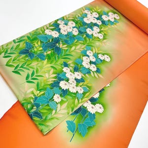Puede incluir: Un textil enrollado con un diseño floral. La tela muestra flores blancas similares a margaritas, hojas de color turquesa y follaje verde sobre un fondo degradado de verde claro a naranja. La parte enrollada es de color naranja sólido.