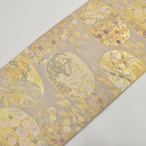 May include: A decorative textile featuring a repeating pattern of oval shapes filled with floral and abstract designs in gold, white, and burgundy on a light grey background. The fabric has a subtle sheen.