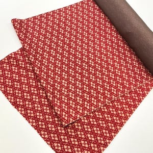 May include: Red fabric with a repeating floral pattern in a lighter shade of red. The fabric is folded and laid out on a white surface, with a rolled-up section of brown fabric visible in the background.