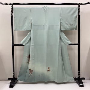 May include: A pale sage green kimono featuring wide sleeves and a central seam. The garment displays delicate, small-scale embroidered figures and floral motifs near the hem and on the left sleeve, set against a smooth, solid-colored fabric.