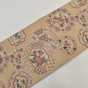 May include: A tan textile featuring a repeating pattern of floral medallions in red, blue, and green. The circular medallions have detailed geometric borders. The fabric, possibly silk, is displayed at an angle, showcasing the design.