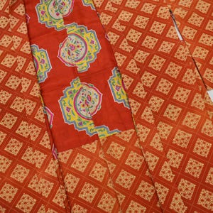 May include: An orange kimono with a repeating diamond pattern and a red panel featuring a floral design with yellow and blue accents. The fabric is folded and displayed against a white background.