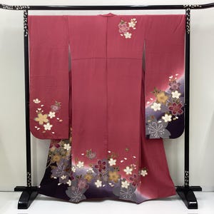 May include: A dusty rose kimono featuring long sleeves and a gradient hem transitioning into deep purple. The garment is decorated with scattered floral motifs in shades of white, gold, and dark pink, with additional blossoms near the shoulder.