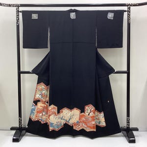 May include: A black kimono with a decorative hem featuring a colorful, traditional Japanese design. The design includes images of birds, trees, and figures in shades of orange, blue, and beige. The kimono is displayed on a black stand.